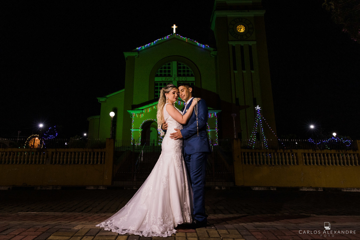casamento de elidi e elias na paróquia do Senhor Bom Jesus matriz de jesuânia MG, fotógrafo de casamentos carlos alexandre foto de três corações lambari mg noivos de frente à igreja ainda com enfeites de natal nessa foto de corpo inteiro