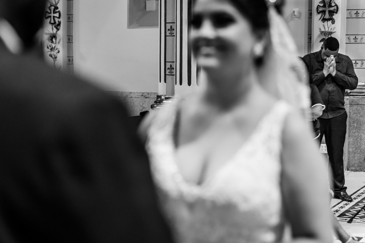 cerimônia de casamento na igreja matriz de três corações mg carlos alexandre fotógrafo amigo do noivo orando ao fundo para que o casal seja abençoado e a noiva continue sorrindo