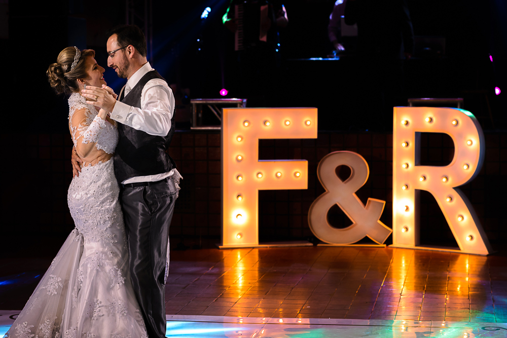 Foto da dança dos noivos e as iniciais em letras caixa com luzes dentro no meio da pista de dança