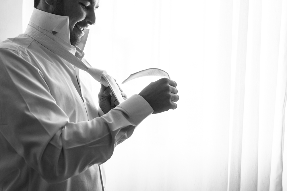Foto do noivo fazendo o nó da gravata em frente a janela do seu quarto