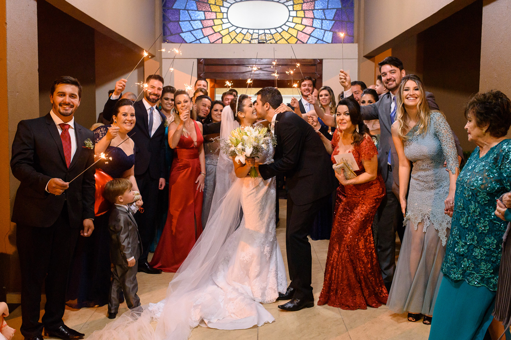 Noivos se beijando na frente da igreja com os padrinhos com sparkles, festejando atrás