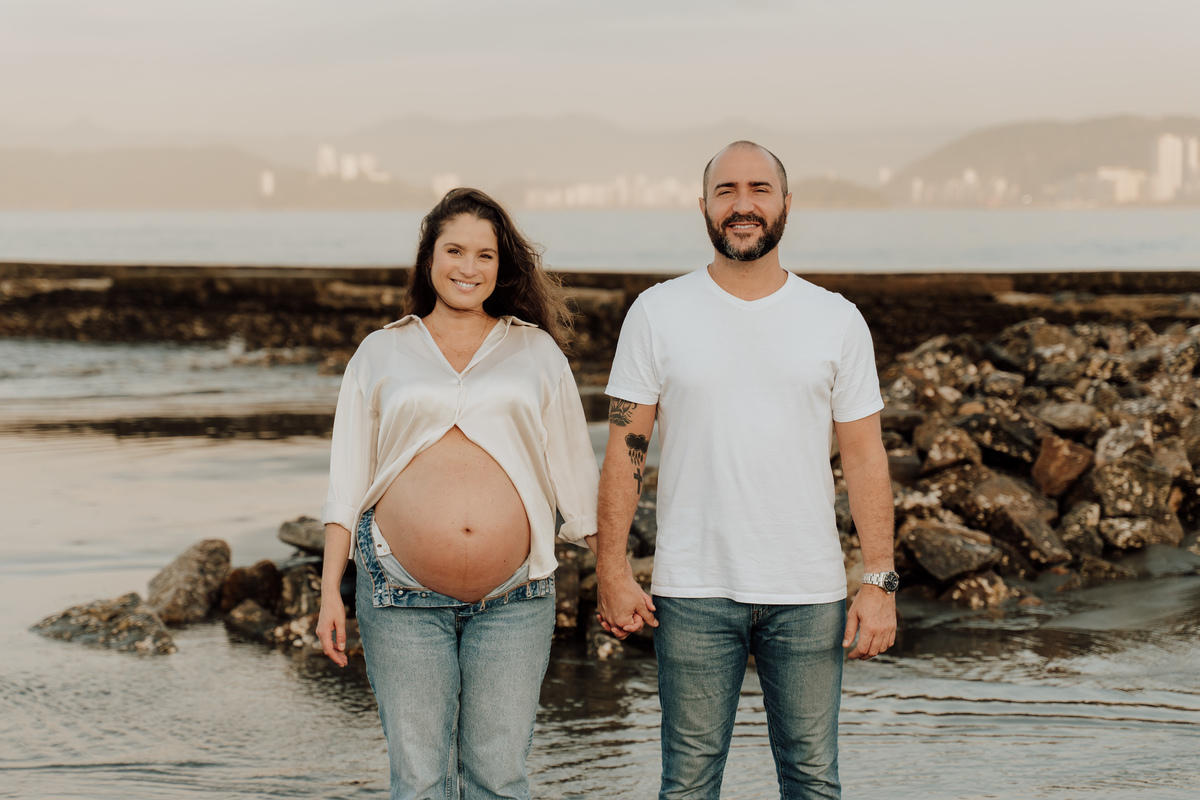 Fotografia de grávida na praia de Santos, usando calça jeans e camisa, capturando a tranquilidade da maternidade com luz natural suave. Foto ideal para ensaios de gestantes que buscam registros emocionantes da gravidez.
