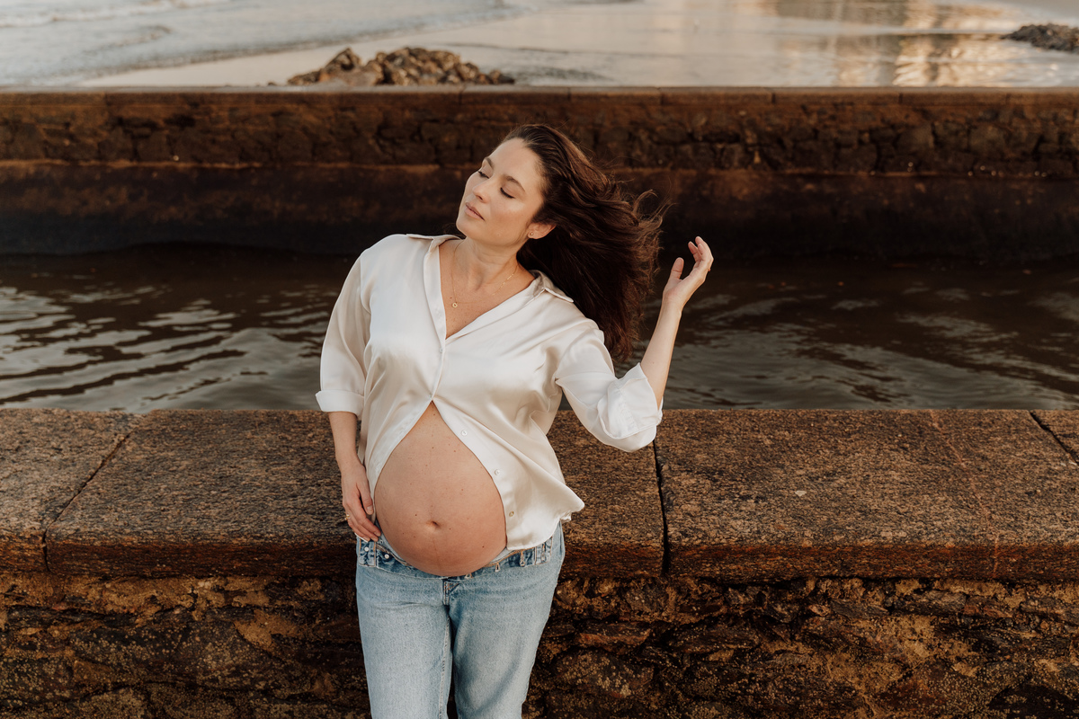 Fotografia de grávida na praia de Santos, usando calça jeans e camisa, capturando a tranquilidade da maternidade com luz natural suave. Foto ideal para ensaios de gestantes que buscam registros emocionantes da gravidez.
