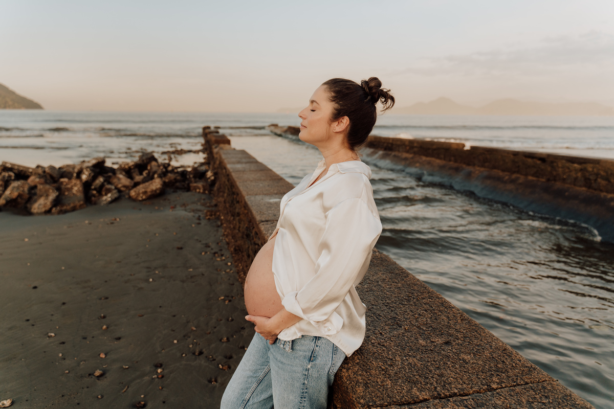 Fotografia de grávida na praia de Santos, usando calça jeans e camisa, capturando a tranquilidade da maternidade com luz natural suave. Foto ideal para ensaios de gestantes que buscam registros emocionantes da gravidez.