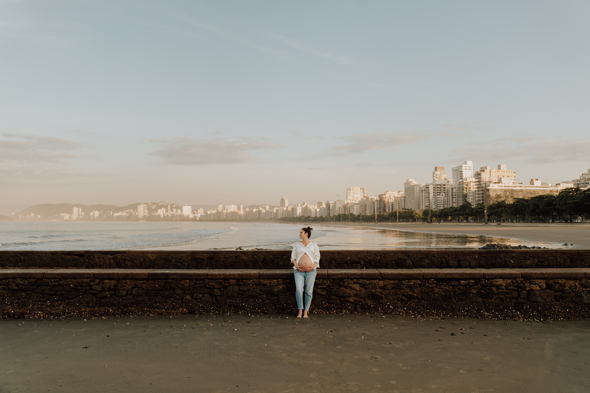 Fotografia de grávida na praia de Santos, usando calça jeans e camisa, capturando a tranquilidade da maternidade com luz natural suave. Foto ideal para ensaios de gestantes que buscam registros emocionantes da gravidez.