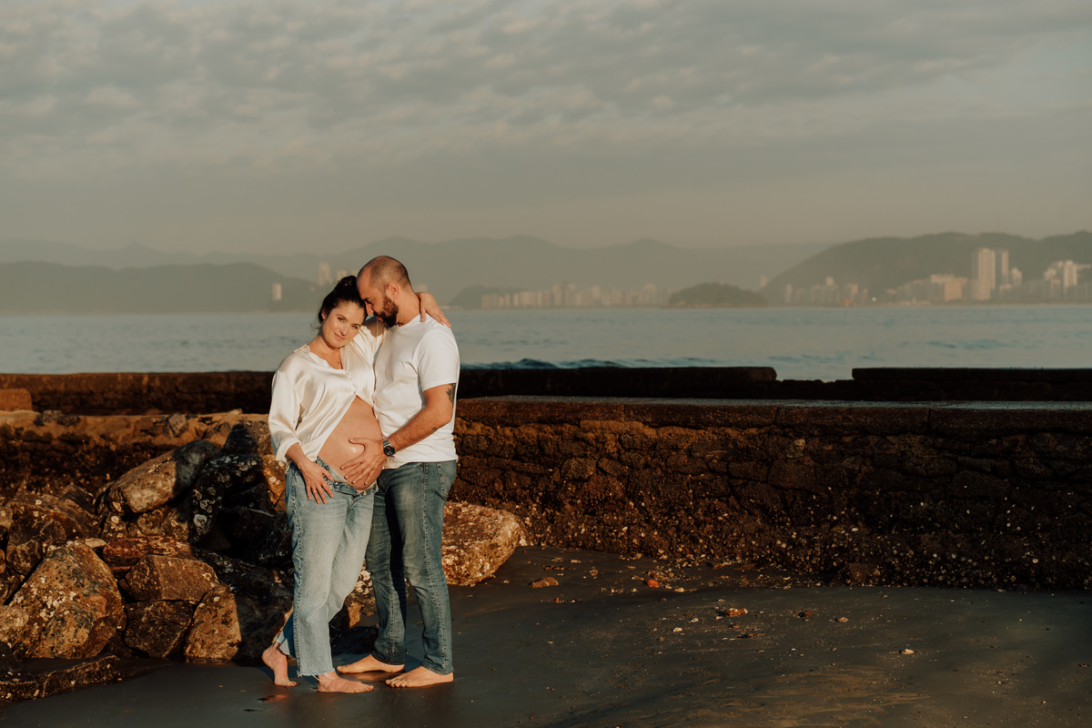 Fotografia de grávida na praia de Santos, usando calça jeans e camisa, capturando a tranquilidade da maternidade com luz natural suave. Foto ideal para ensaios de gestantes que buscam registros emocionantes da gravidez.
