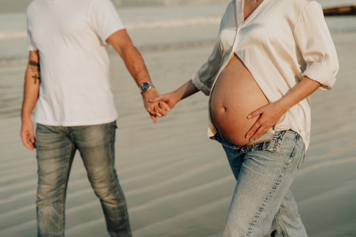 Fotografia de grávida na praia de Santos, usando calça jeans e camisa, capturando a tranquilidade da maternidade com luz natural suave. Foto ideal para ensaios de gestantes que buscam registros emocionantes da gravidez.