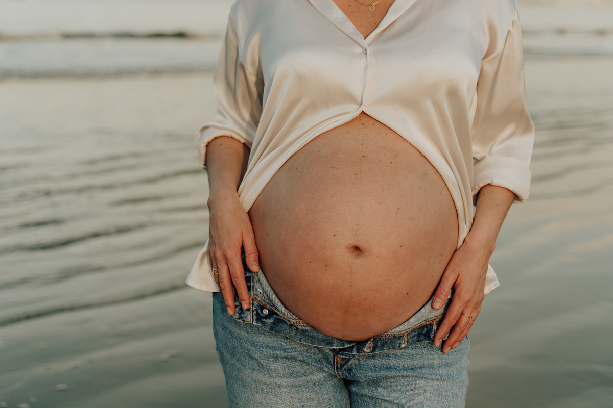 Fotografia de grávida na praia de Santos, usando calça jeans e camisa, capturando a tranquilidade da maternidade com luz natural suave. Foto ideal para ensaios de gestantes que buscam registros emocionantes da gravidez.