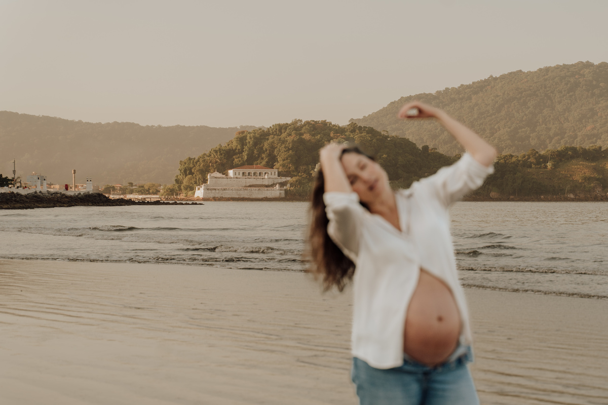 Fotografia de grávida na praia de Santos, usando calça jeans e camisa, capturando a tranquilidade da maternidade com luz natural suave. Foto ideal para ensaios de gestantes que buscam registros emocionantes da gravidez.