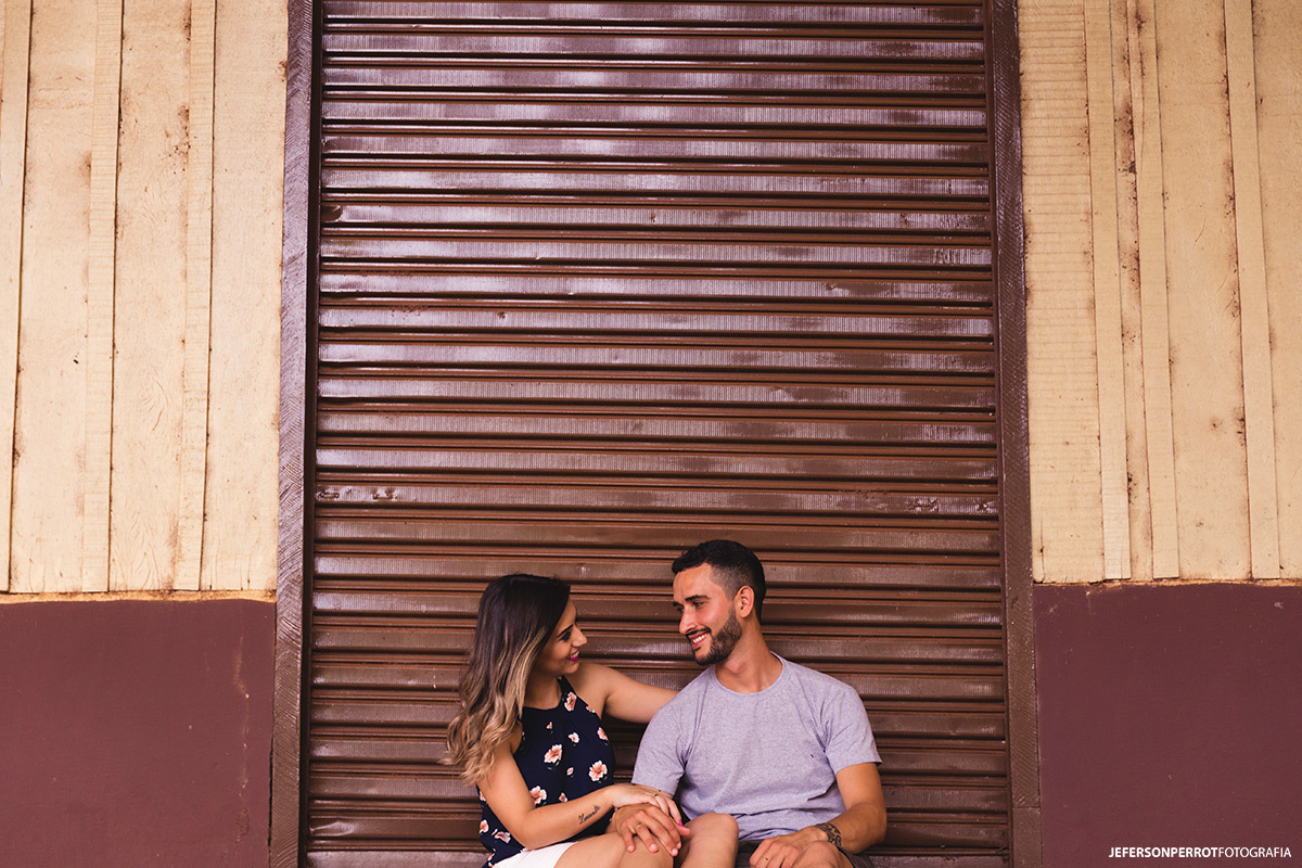 casal sentado em frente a uma porta de ferro rústica em ensaio pré-casamento em mandaguacu, no parana