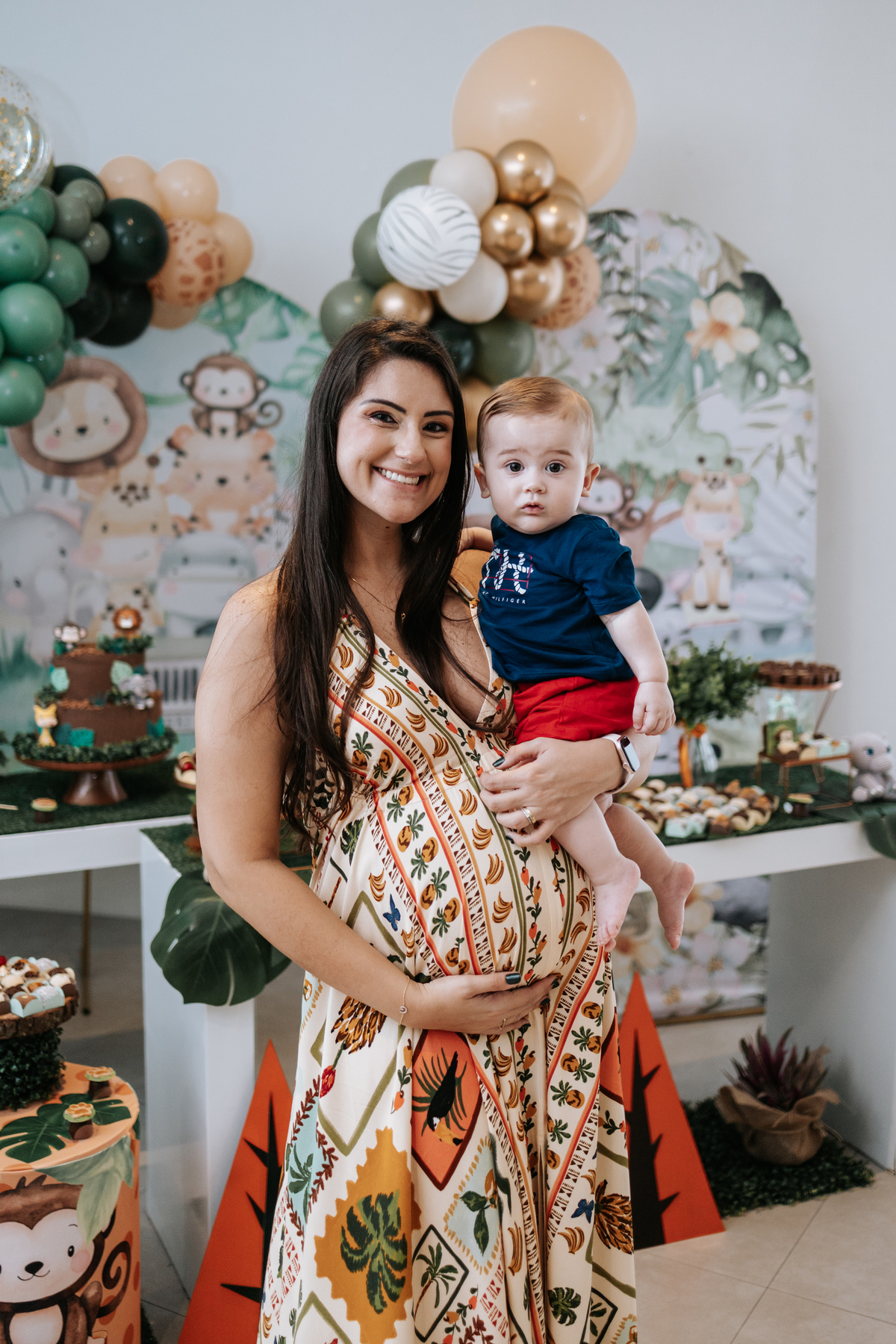 Foto da gestante segurando uma criança no colo próxima à mesa do bolo.