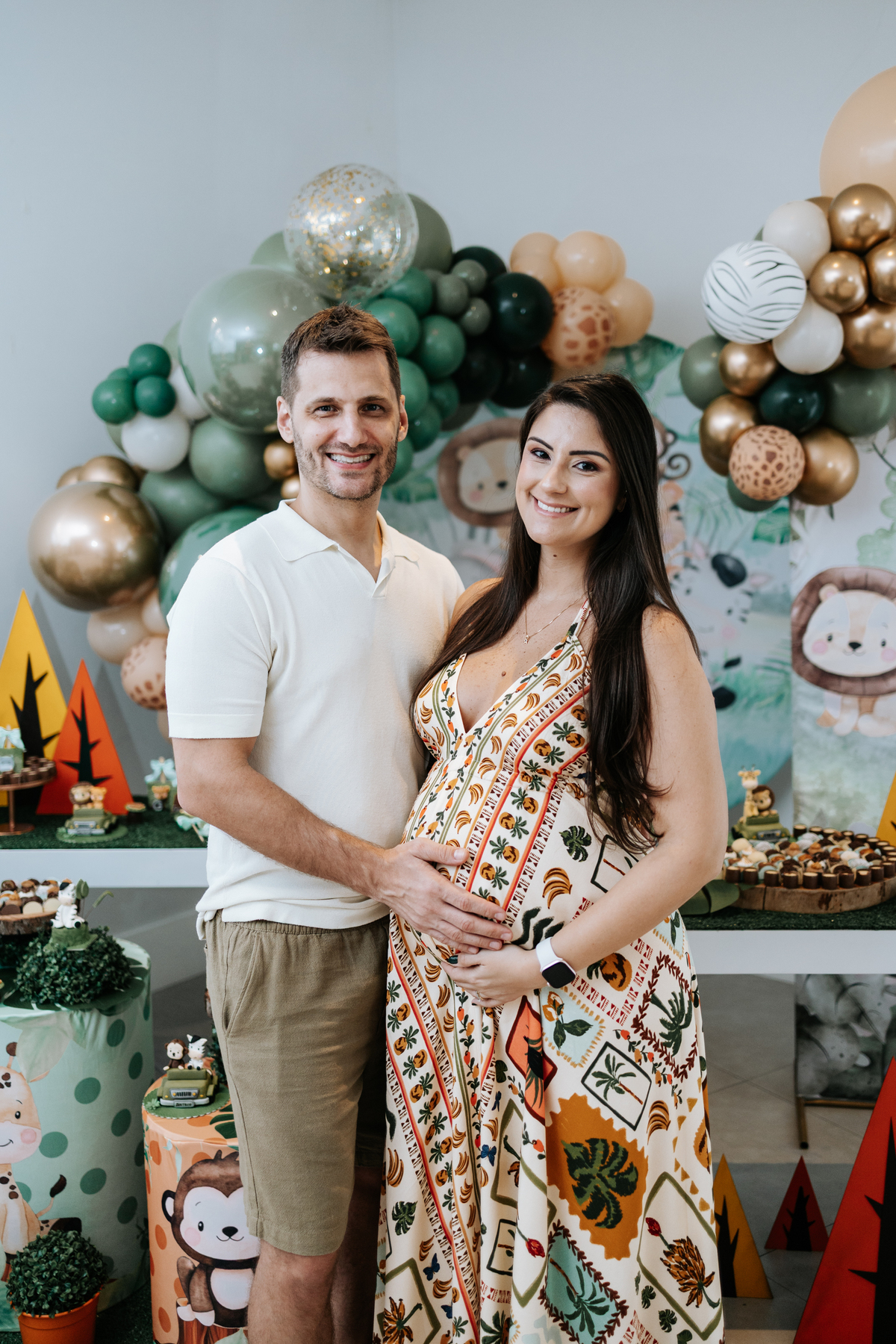 Foto do casal sorridente com as mãos na barriga em frente à decoração.