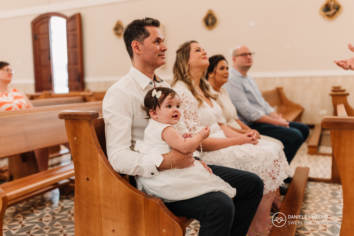 Pais segurando a bebê nos braços com carinho, durante a cerimônia de batismo na Igreja do Socorro.