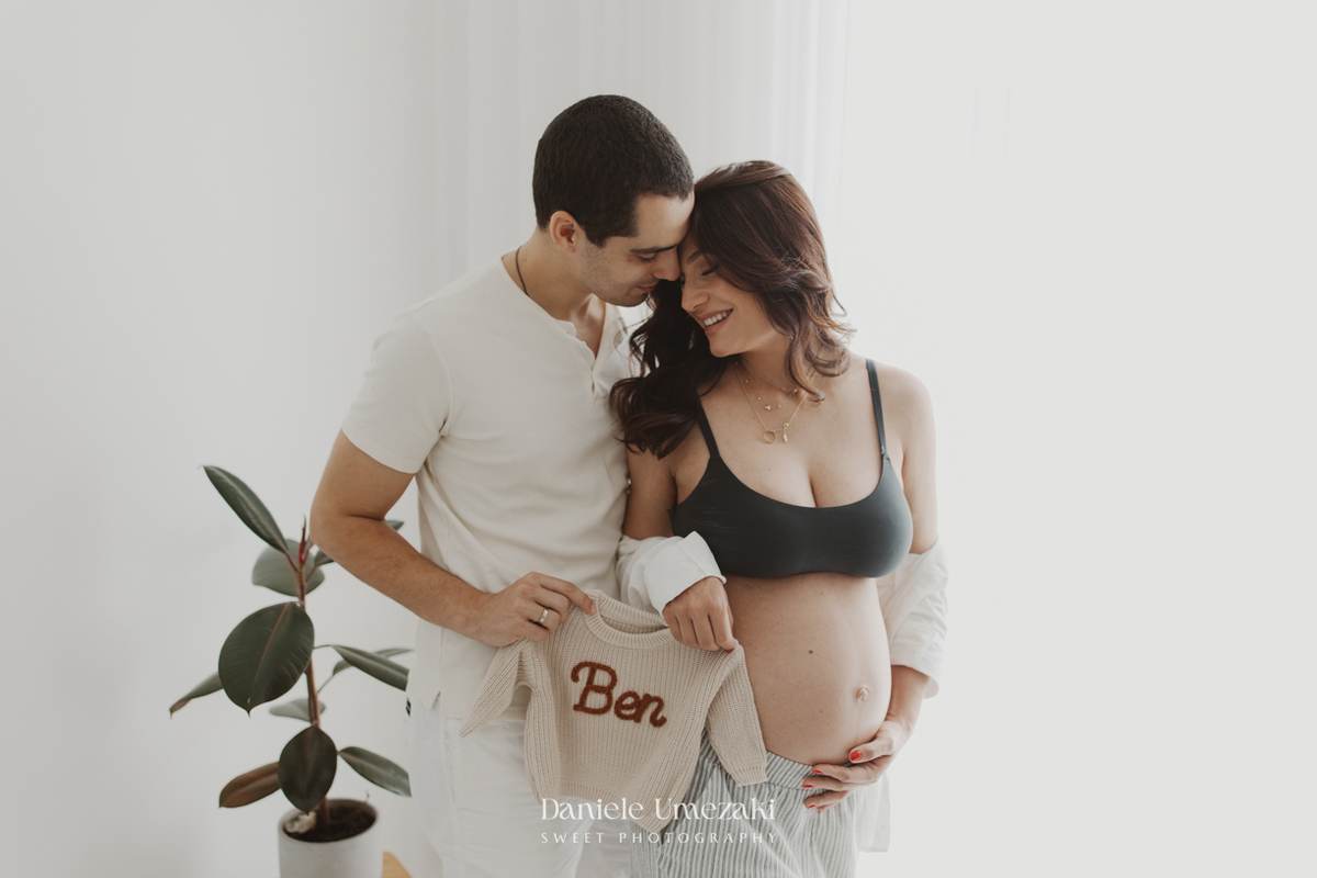 Ensaio gestante em estúdio em Mogi das Cruzes com detalhe da barriga e roupinha do bebê Ben, em um registro delicado e afetivo.