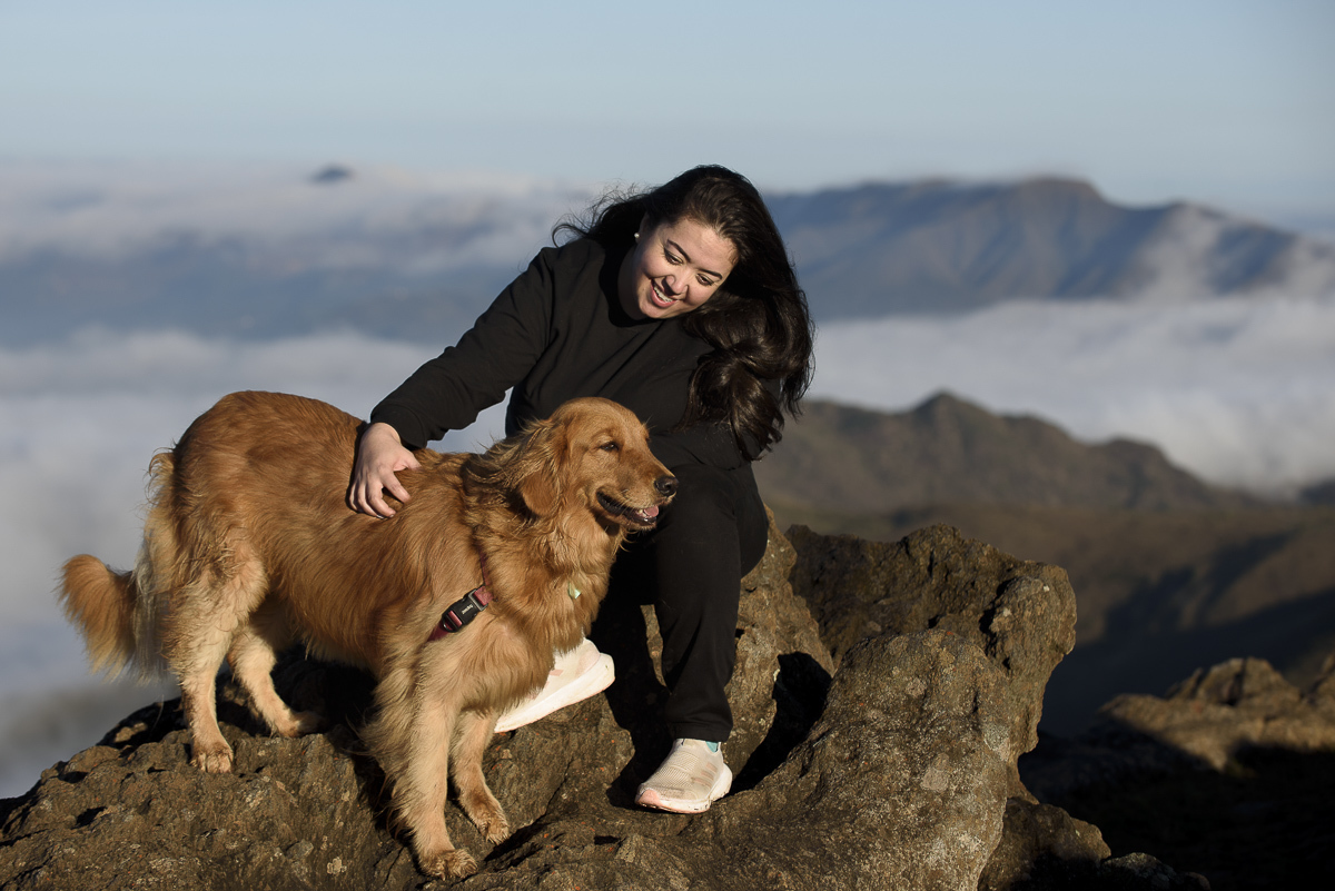 Moça sentanda na pedra olhando em direcão ao seu golden retriever e sorrindo levemente. Ao fundo nunven e a vista da montanha