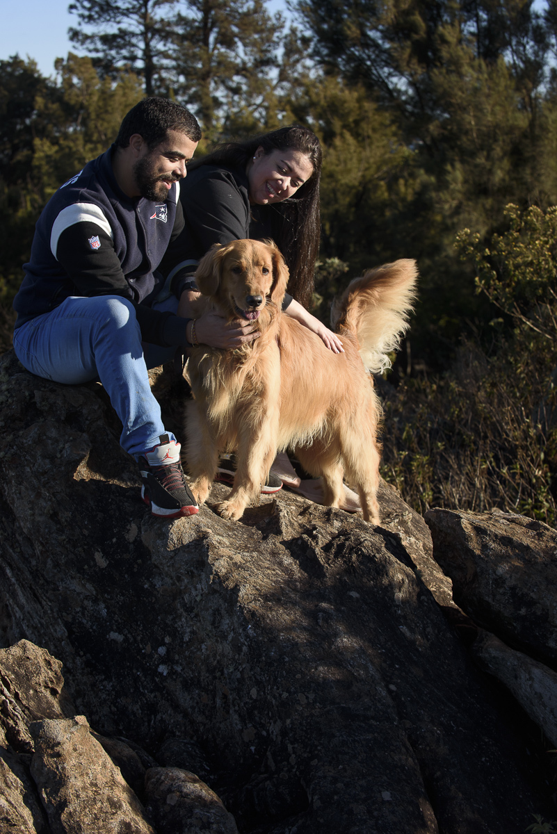 Numa foto mais fechada o Casal está numa pedra mais iclinada e alta com sua golden retriever que está com rabo levantado. Todos olham para frente e sorriem 