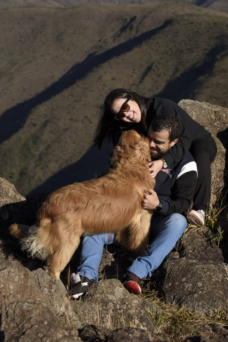 Uma foto mais fechada de um momento de descontração entre o casal e a golden retriever. A moça apaioda na pedra , levemente abraçada no rapaz que está sentado numa pedra mais baixa. Ambos fazem fazem carinho na golden