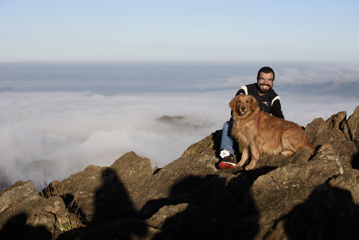 rapaz sentando na pedra com seu golden retriever. ambos olham para frente . Ao fundo nuvens baixas de uma manhã linda e fria