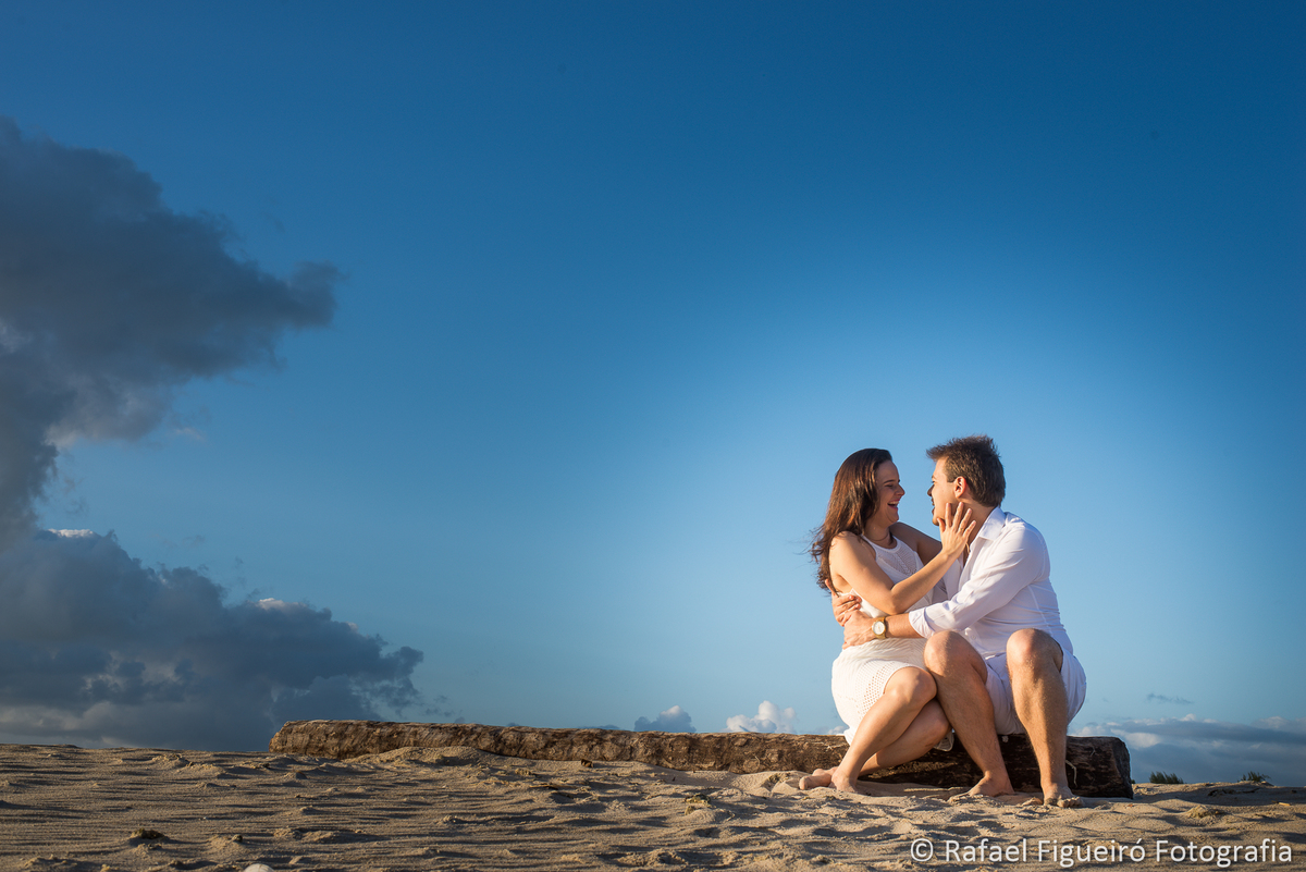 casal namorando sorindo sentado tronco de coqueiro praia areia céu azul