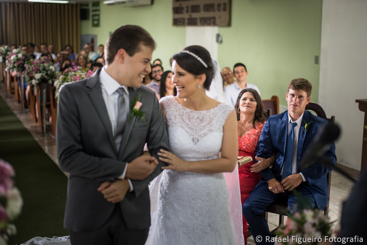 noivos se olhando e sorrindo cerimonia de casamento mãe e irmão sorrindo ao fundo sentados