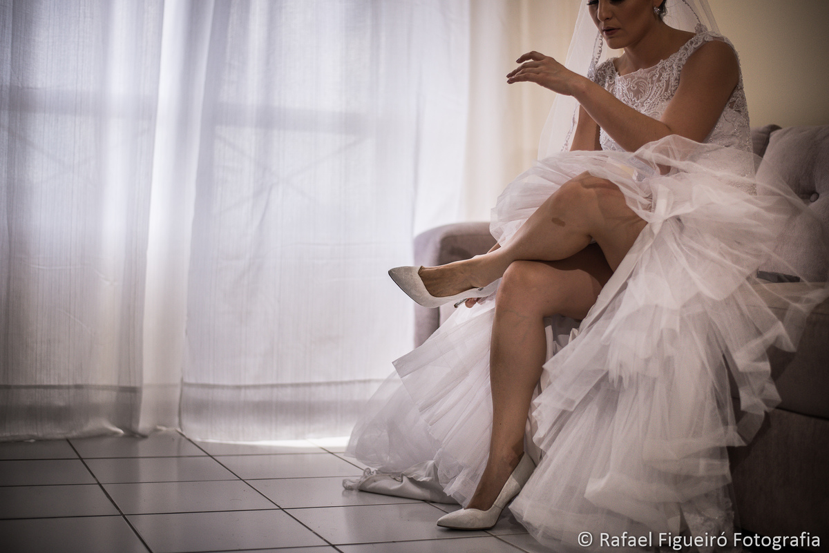 noiva sentada no sofá calçando sapatos pernas cruzadas cortina branca ao fundo