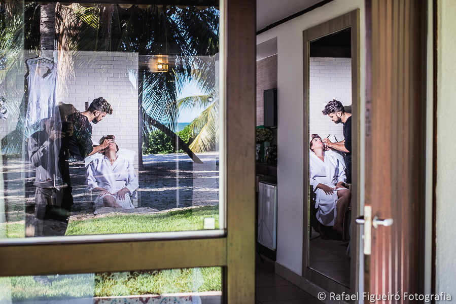 making of da noiva sendo maquiada com reflexos de espelho e paisagem deslumbrante de coqueiros e mar refletidos no vidro da janela fotografado por Rafael Figueiró, fotógrafo de casamentos em Recife-PE
