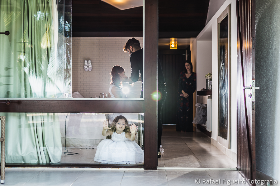 daminha de honra brincando com o rosto e mãos coladas no vidro da janela noiva é maquiada ao fundo fotografado por Rafael Figueiró fotógrafo de casamentos em Recife-PE pernambuco