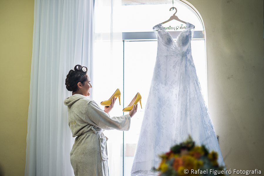 Noiva admira os Sapatos amarelos segurando em mãos vestido pendurado na janela fotografado por Rafael Figueiró melhor fotógrafo de casamentos de Recife-PE