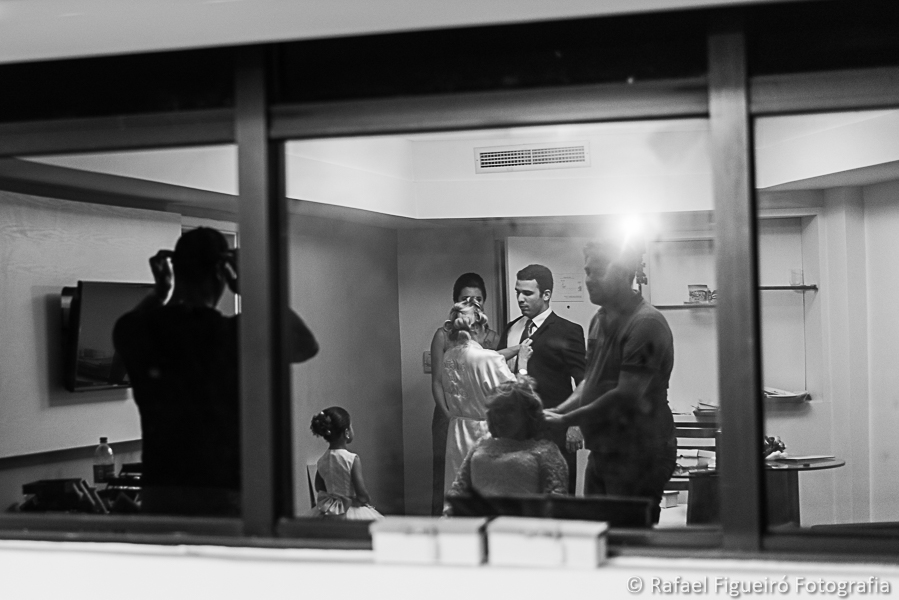 imagem refletida no vidro da janela noiva dando nó da gravata de padrinho fotografado por rafael figueiró melhor fotógrafo de casamentos em recife-pe