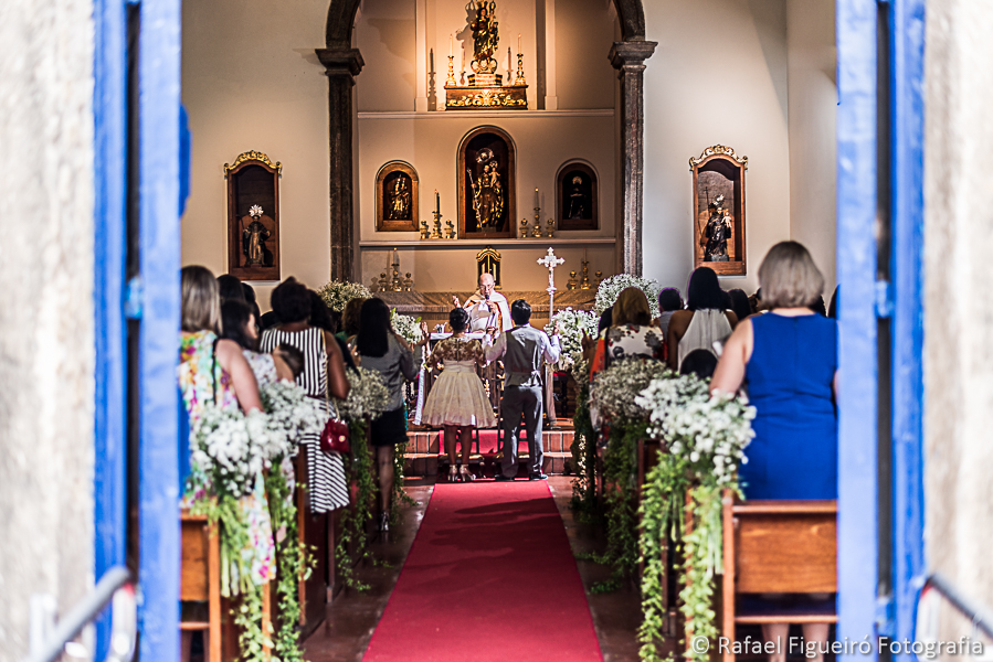 Casal de costas no altar com padre ao fundo visto de fora da igreja fotografado por Rafael Figueiró melhor fotógrafo de casamentos de Recife-PE