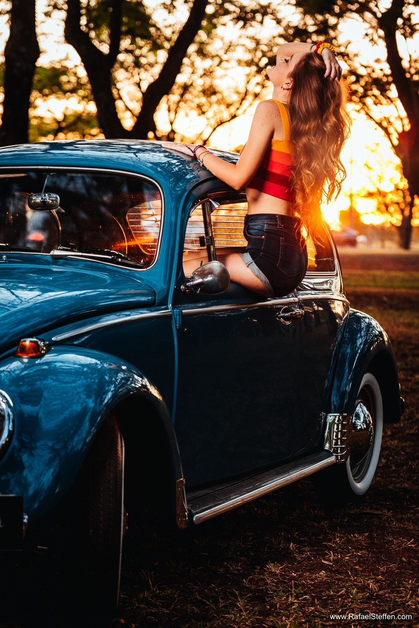 Foto do Ensaio de quinze anos da Ester sentada na janela do fusca azul no Pôr do sol com as mãos nos cabelos olhando para cima fotografado por Rafael Steffen Fotografia.