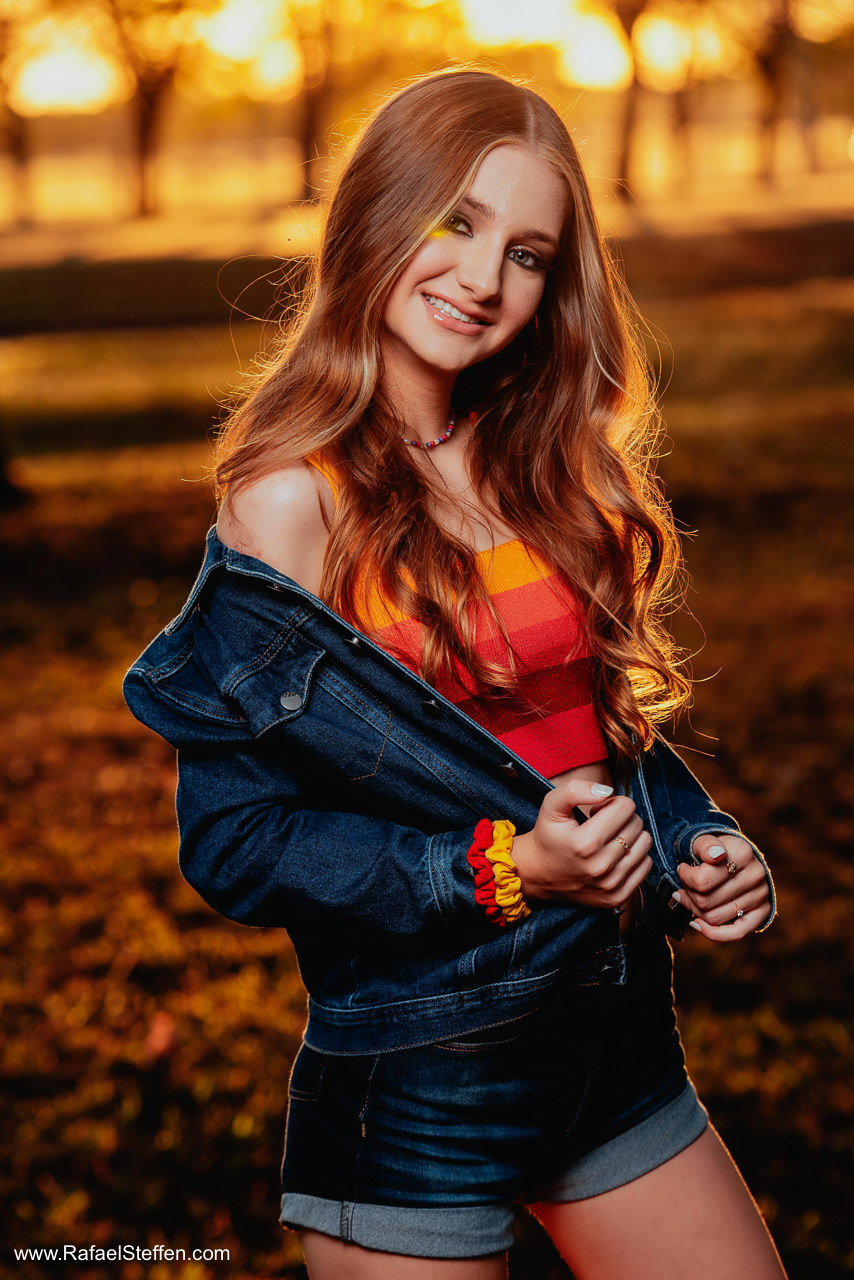 Foto do ensaio de quinze anos da Ester olhando para baixo com jaqueta jeans sorrindo no pôr do sol em Brasília fotografado por Rafael Steffen Fotografia.