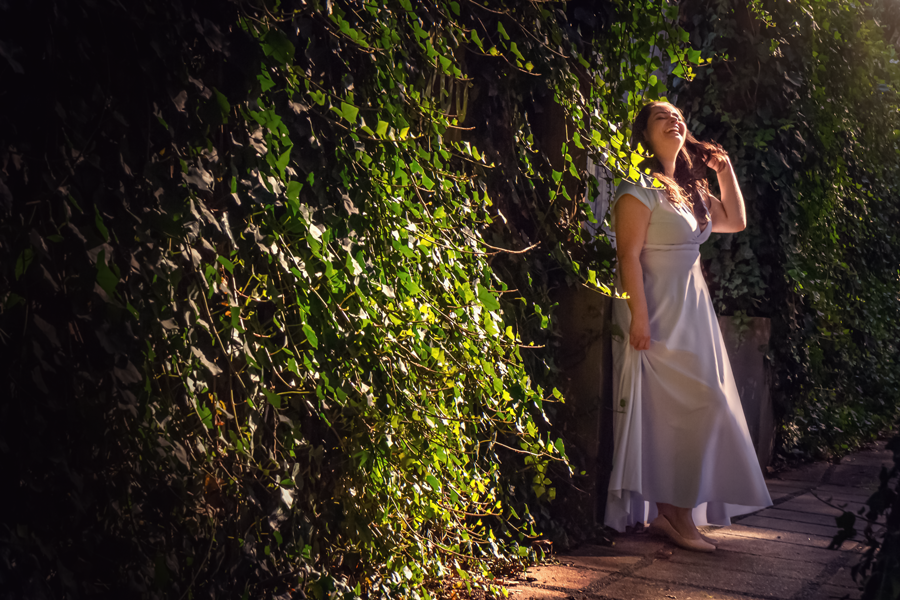 garota sorrindo em frente a um muro coberto de plantas verdes-luzes e sombras-mulher segura vestido o longo branco-garota passa a mao no cabelo-garota feliz-fotos em porto alegre