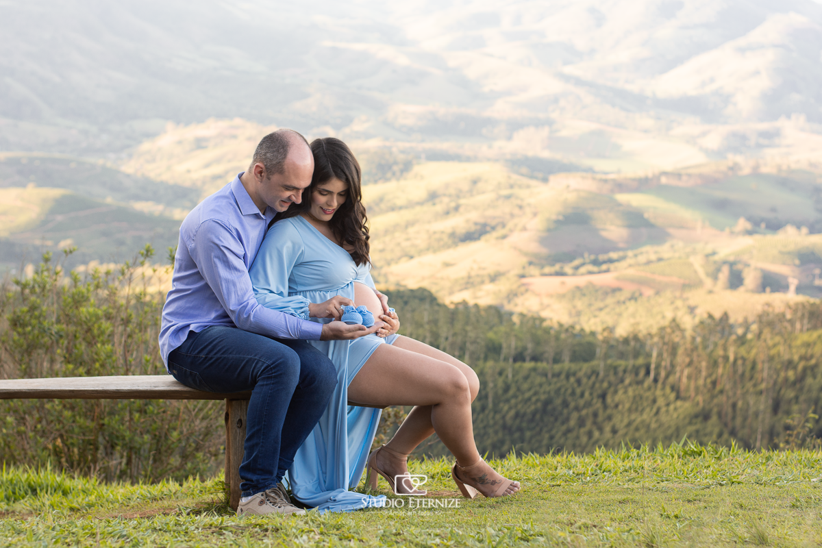 Ensaio fotográfico de gestante da Juliana e do Lucas em um lindo local externo em Poços de caldas, sentados em um banquinho de madeira na Rampa de Paraglider estão admirando o Miguel e seu sapatinho de linha azul