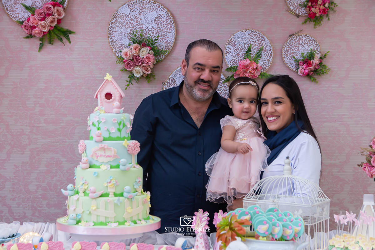 Fotografia da aniversariante Giovanna  com seus pais na mesa do bolo.