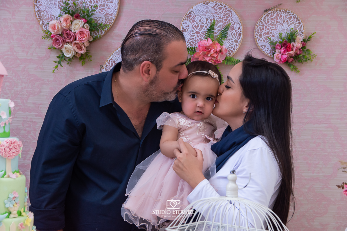Fotografia da aniversariante Giovanna  com seus pais na mesa do bolo, sanduiche de beijo.