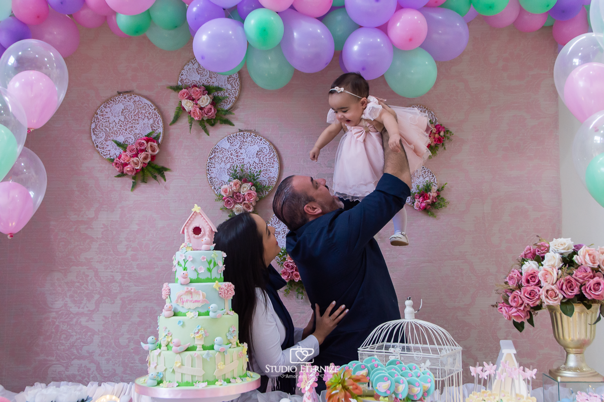 Fotografia da aniversariante Giovanna  com seus pais na mesa do bolo.