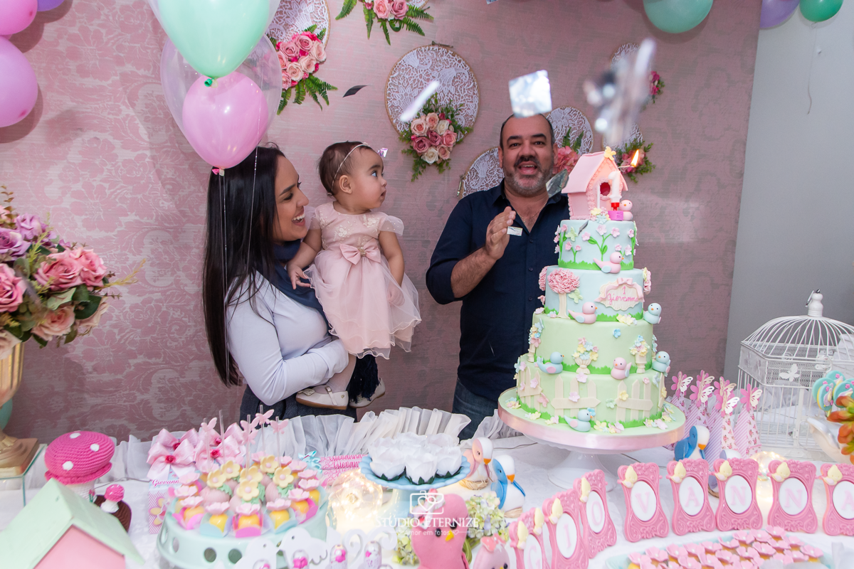  Fotografia da aniversariante e seus pais na mesa do bolo cantando parabéns.