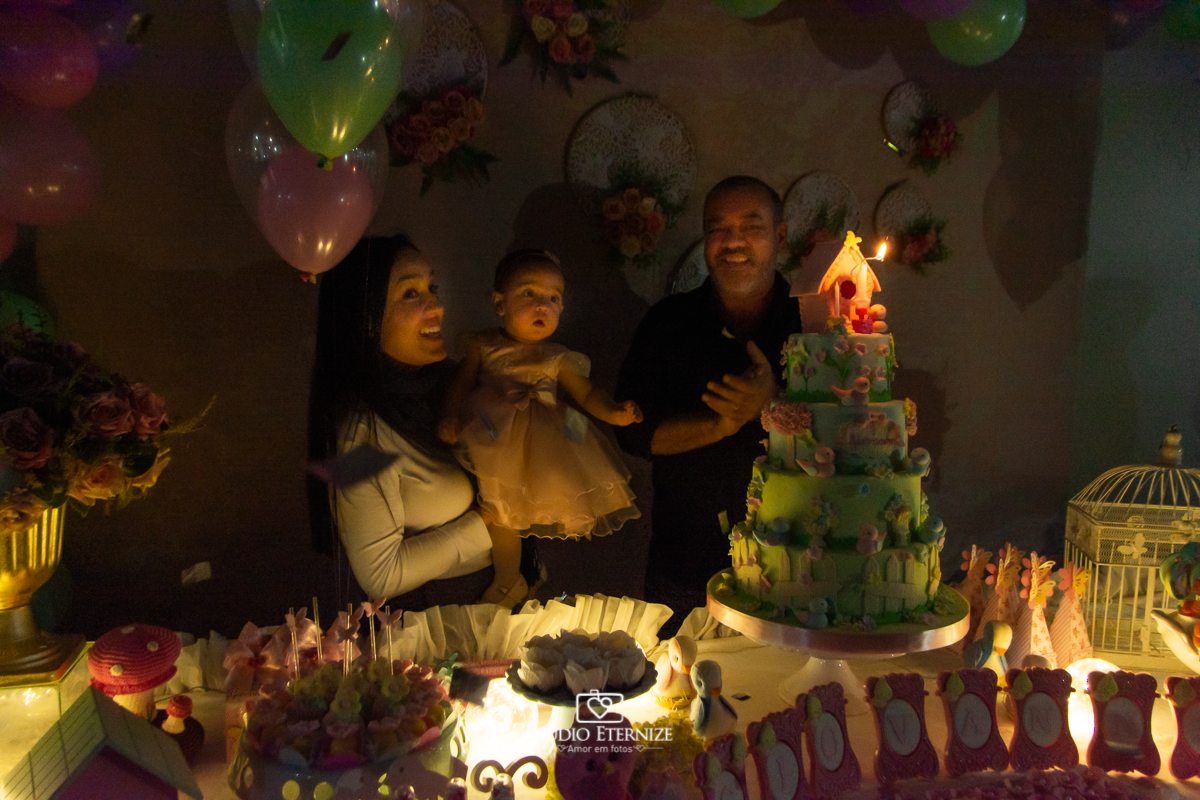  Fotografia da aniversariante e seus pais na mesa do bolo cantando parabéns.