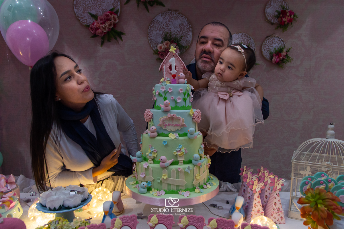  Fotografia da aniversariante e seus pais na mesa do bolo soprando a velinha.