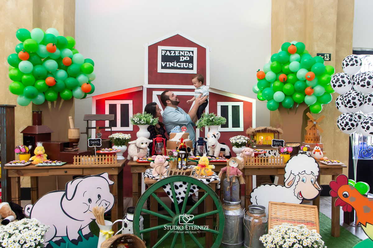Fotografia de aniversario infantil no tema fazendinha realizado em Poços de Caldas. Pais brincando de jogar aniversariante para cima, atrás da mesa do bolo.