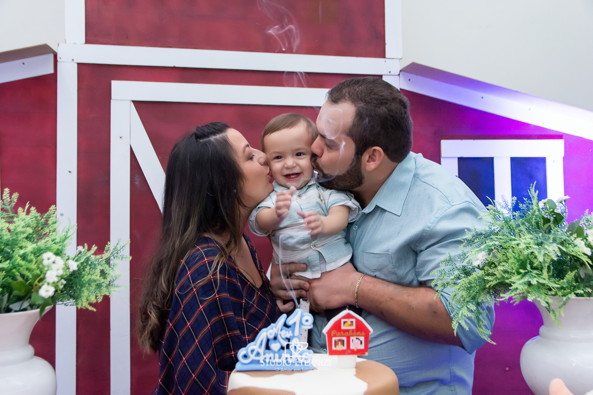 Fotografia de aniversario infantil no tema fazendinha realizado em Poços de Caldas. Pais fazendo sanduíche de beijo no aniversariante na mesa do bolo.