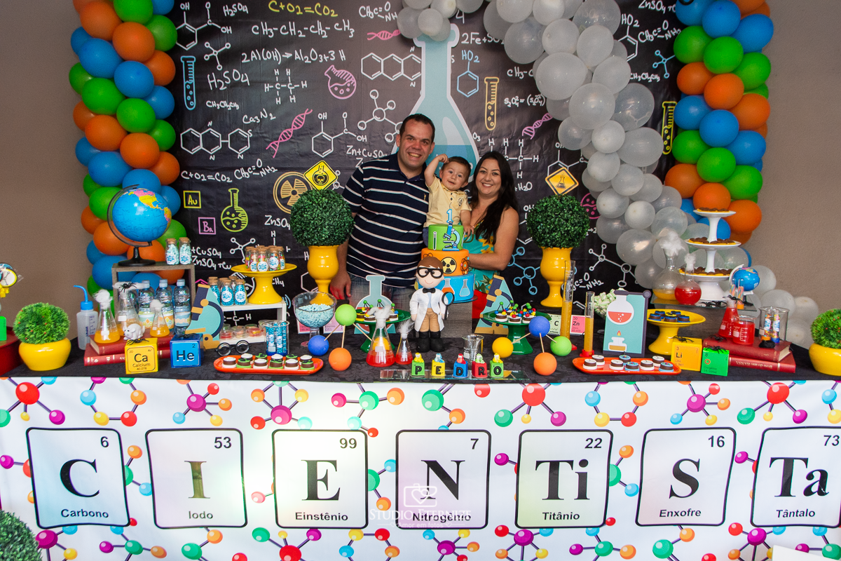 Fotografia de aniversário infantil realizado pelo Studio Eternize em Poços de Caldas. Tema da festa cientista. Aniversariante e seus pais posando para a foto na mesa do bolo.