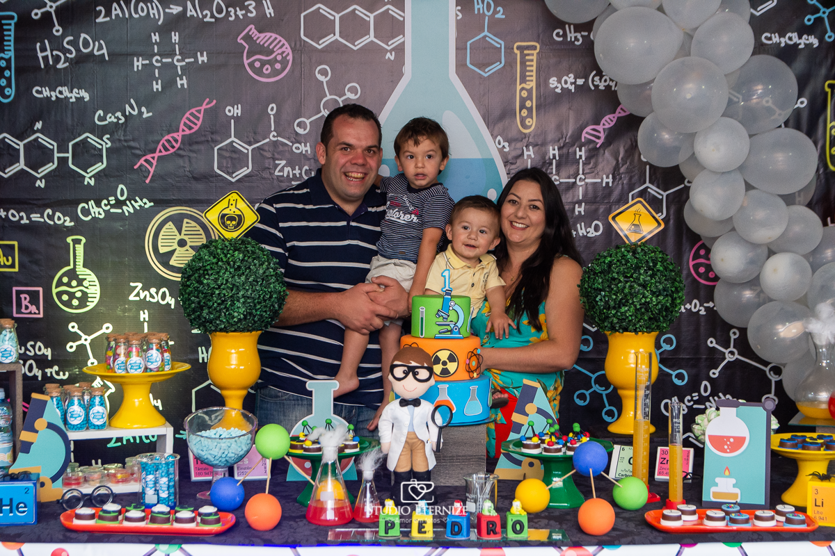 Fotografia de aniversário infantil realizado pelo Studio Eternize em Poços de Caldas. Tema da festa cientista. Aniversariante, seu irmão e seus pais posando para a foto na mesa do bolo.