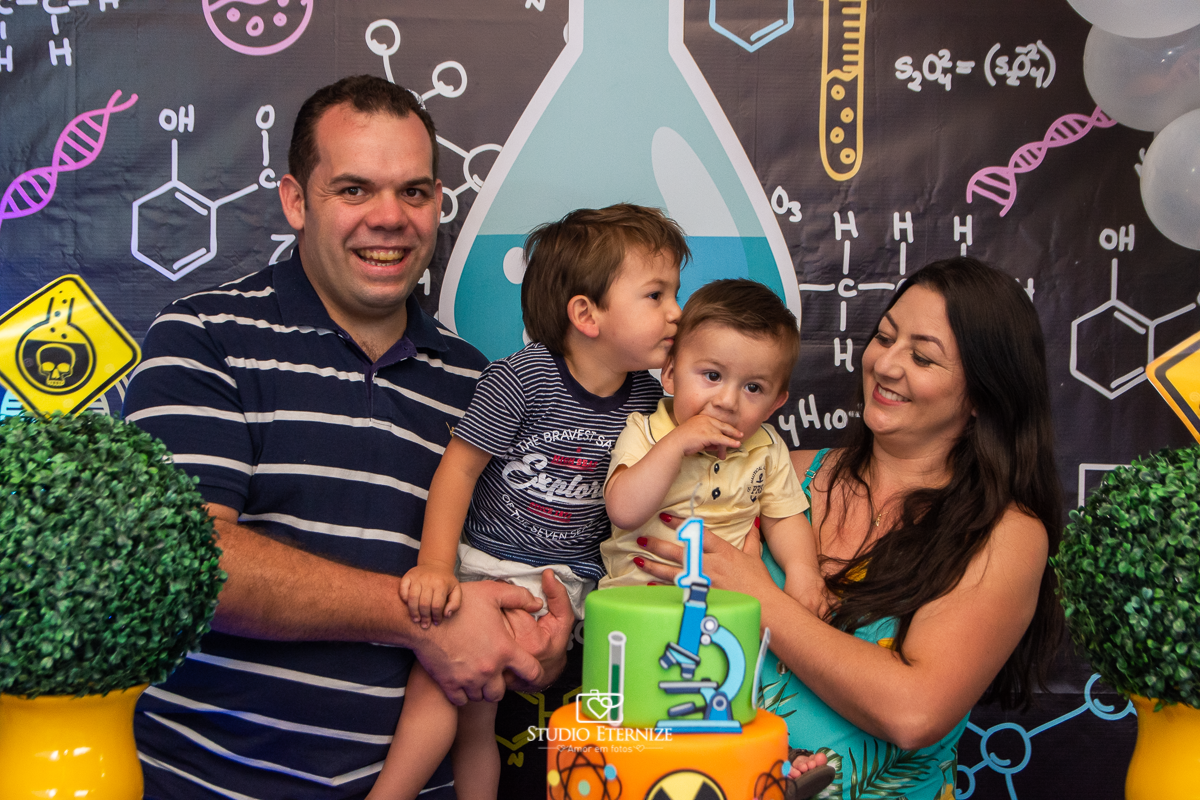 Fotografia de aniversário infantil realizado pelo Studio Eternize em Poços de Caldas. Tema da festa cientista. Aniversariante recebendo um beijo do seu irmão no colo dos seus pais posando para a foto na mesa do bolo.