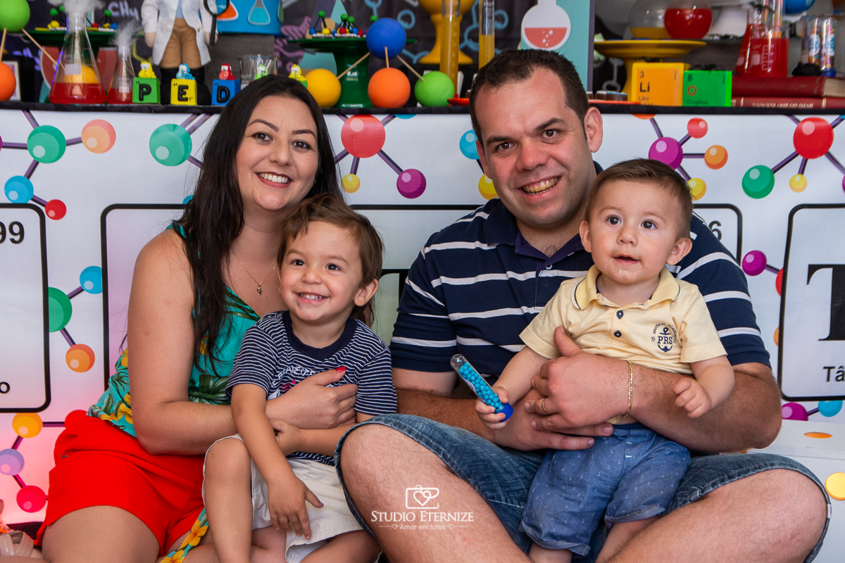 Fotografia de aniversário infantil realizado pelo Studio Eternize em Poços de Caldas. Tema da festa cientista. Aniversariante, seu irmão e seus pais posando para a foto em frente a mesa do bolo.