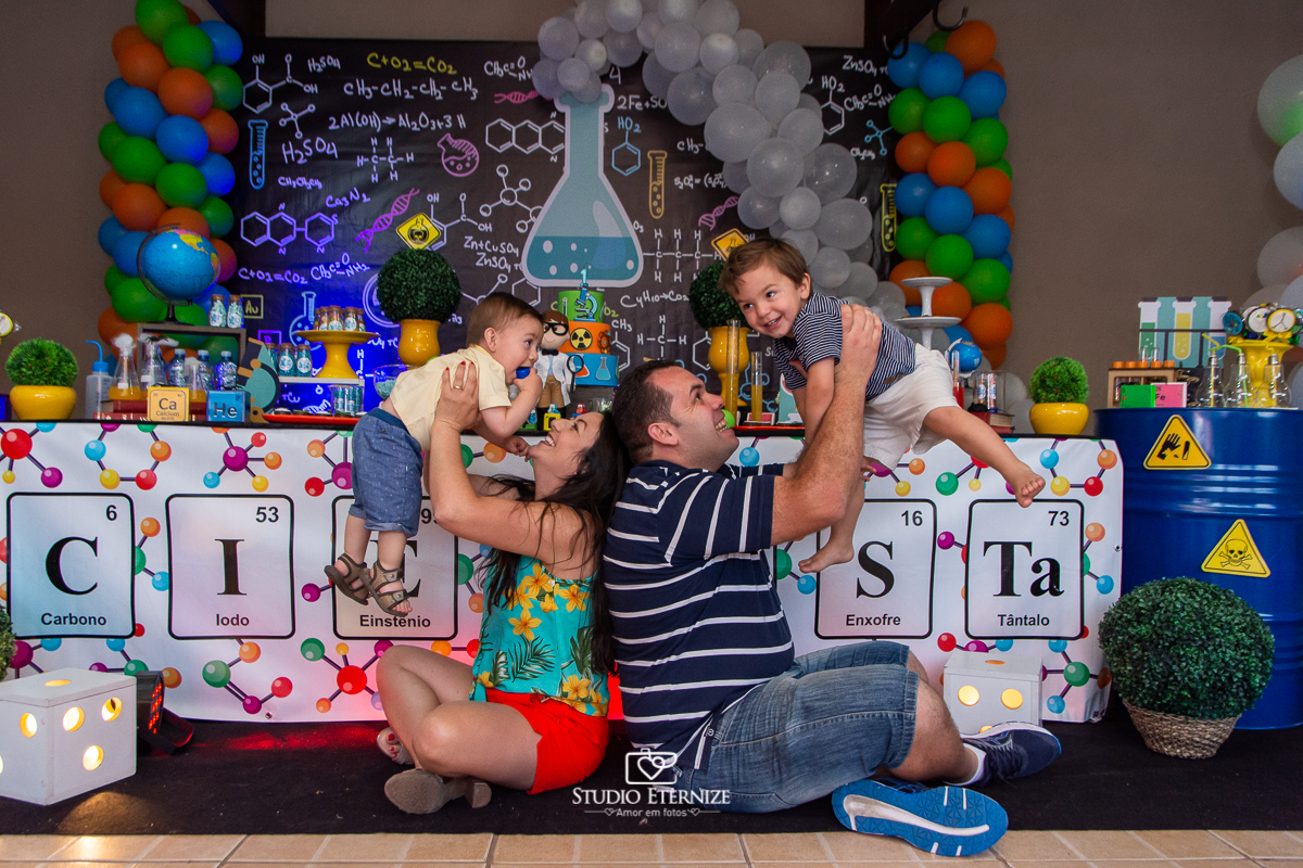 Fotografia de aniversário infantil realizado pelo Studio Eternize em Poços de Caldas. Tema da festa cientista. Aniversariante e seu irmão sendo levantados por seus pais em frente a mesa do bolo.