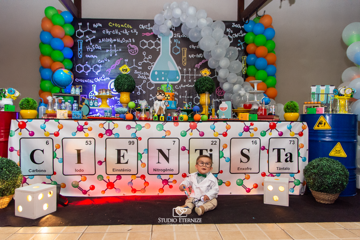 Fotografia de aniversário infantil realizado pelo Studio Eternize em Poços de Caldas. Tema da festa cientista. Aniversariante de óculos posando para a foto em frente a mesa do bolo, em um plano geral.