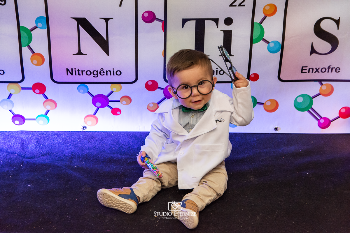 Fotografia de aniversário infantil realizado pelo Studio Eternize em Poços de Caldas. Tema da festa cientista. Aniversariante de óculos posando para a foto em frente a mesa do bolo.