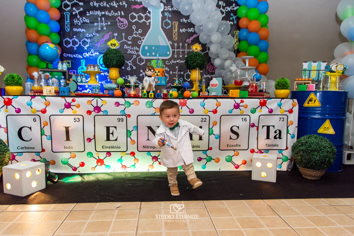 Fotografia de aniversário infantil realizado pelo Studio Eternize em Poços de Caldas. Tema da festa cientista. Aniversariante andando em frente a mesa do bolo, em um plano geral.